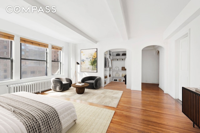 bedroom featuring radiator heating unit, arched walkways, beam ceiling, and wood finished floors