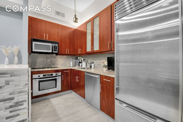 kitchen featuring stainless steel appliances, backsplash, glass insert cabinets, hanging light fixtures, and light stone countertops