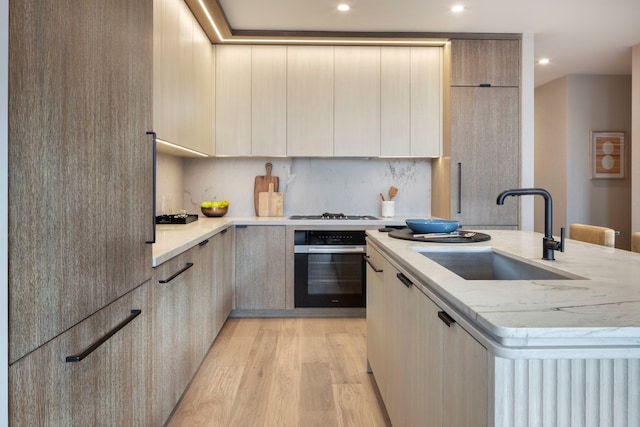 kitchen with oven, a sink, modern cabinets, a center island with sink, and light wood-style floors
