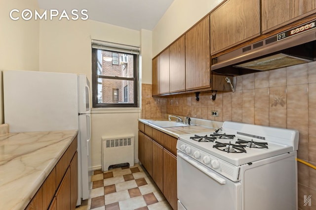 kitchen featuring brown cabinets, a sink, radiator heating unit, under cabinet range hood, and white appliances