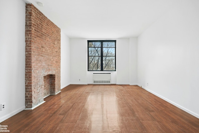 unfurnished living room featuring radiator heating unit, wood finished floors, a brick fireplace, and baseboards