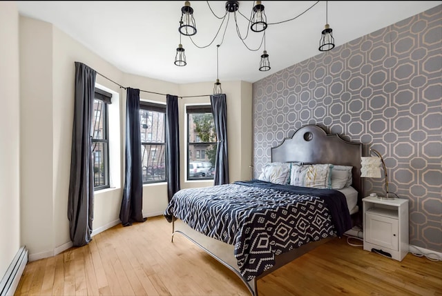 bedroom with wood finished floors, wallpapered walls, baseboard heating, baseboards, and an accent wall