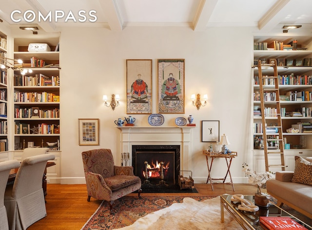 living area with beamed ceiling, wood finished floors, ornamental molding, a fireplace with flush hearth, and bookshelves