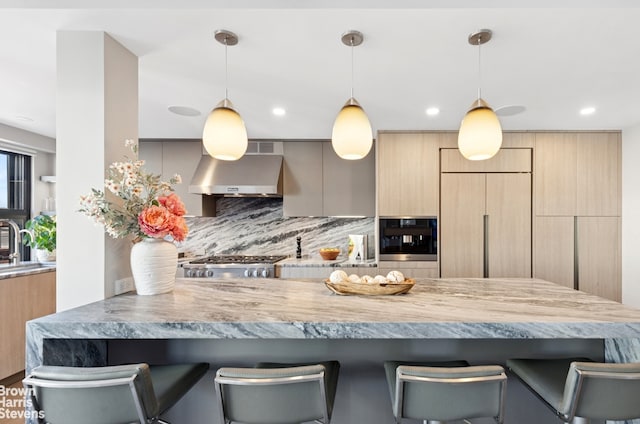 kitchen featuring light brown cabinets, oven, a kitchen breakfast bar, wall chimney range hood, and tasteful backsplash