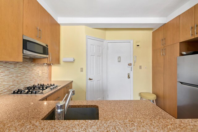 kitchen featuring stainless steel appliances, light stone counters, tasteful backsplash, a sink, and brown cabinetry