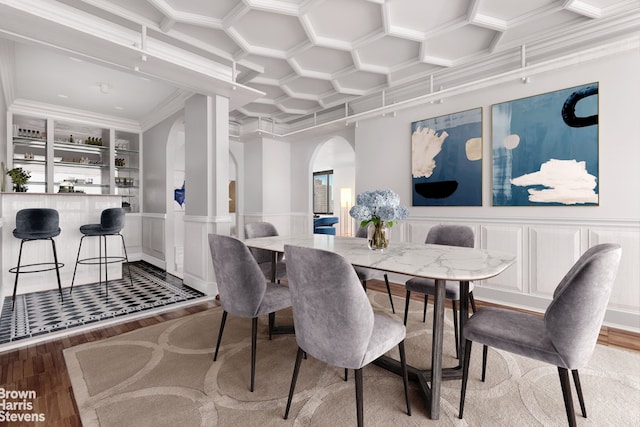 dining room featuring arched walkways, wood finished floors, coffered ceiling, ornamental molding, and wainscoting