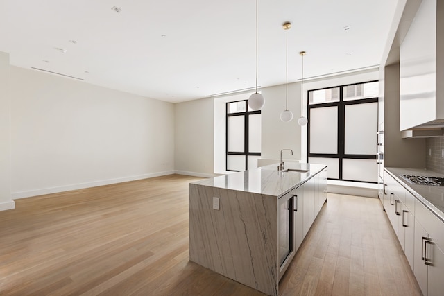 kitchen featuring a sink, light wood-style floors, wall chimney exhaust hood, a center island with sink, and modern cabinets