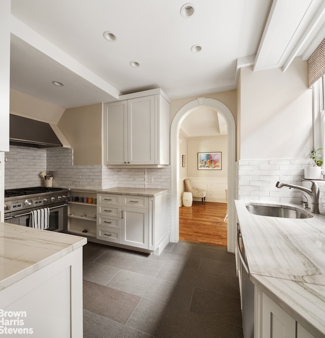 kitchen with stainless steel appliances, a sink, wall chimney exhaust hood, light stone counters, and a wainscoted wall