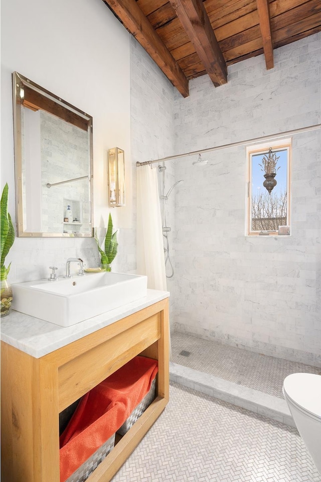 full bath featuring a tile shower, beamed ceiling, vanity, toilet, and wooden ceiling