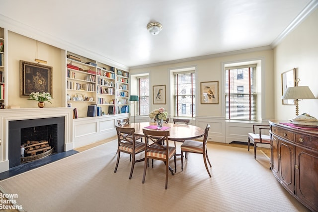 dining space featuring built in shelves, a decorative wall, crown molding, wainscoting, and a fireplace with flush hearth