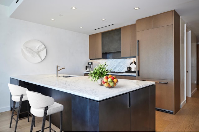 kitchen featuring a sink, light wood finished floors, tasteful backsplash, light stone counters, and a kitchen breakfast bar