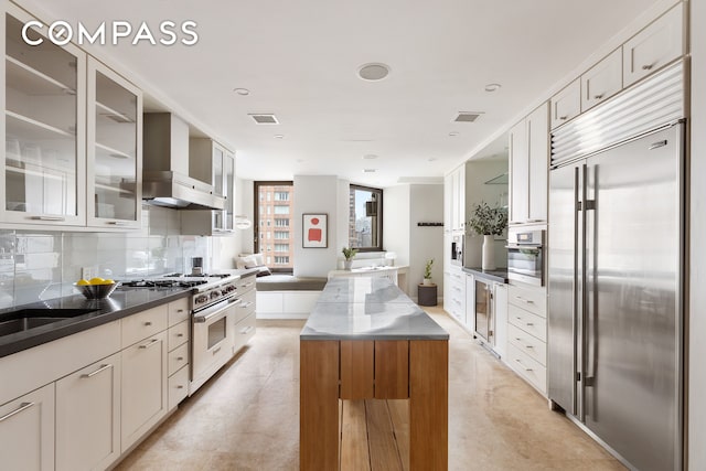 180 E 79th St Unit 14G, New York City, NY 10075 - kitchen featuring premium appliances, wall chimney exhaust hood, tasteful backsplash, glass insert cabinets, and white cabinets