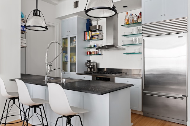 kitchen featuring stainless steel appliances, open shelves, dark countertops, wall chimney exhaust hood, and a breakfast bar area