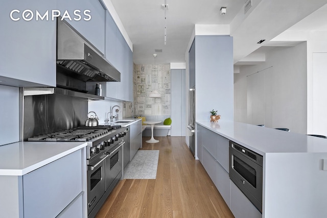 kitchen with high end appliances, modern cabinets, wall chimney exhaust hood, light wood finished floors, and light countertops