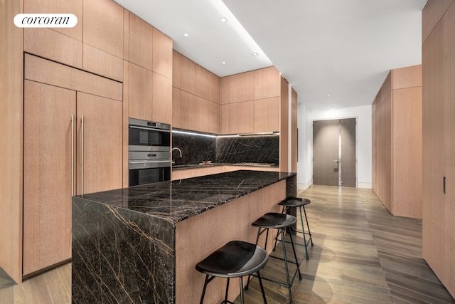 kitchen featuring light wood-style floors, dark stone counters, modern cabinets, double oven, and a kitchen breakfast bar
