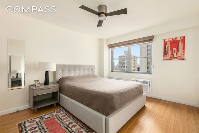 bedroom with baseboards, light wood-type flooring, a wall unit AC, and a ceiling fan