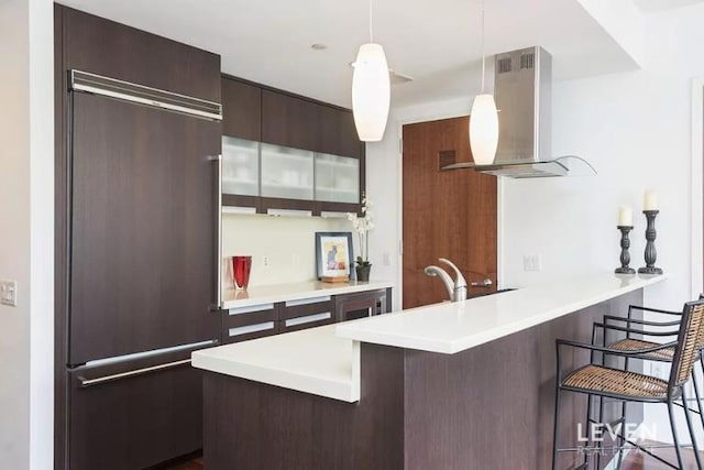 kitchen featuring a peninsula, paneled built in fridge, a kitchen bar, light countertops, and wall chimney exhaust hood