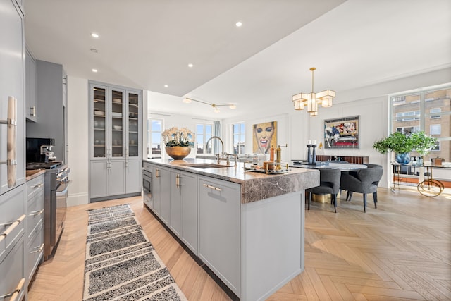 kitchen with a sink, range with two ovens, gray cabinetry, a center island with sink, and a chandelier