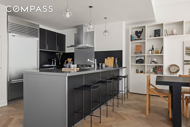 kitchen with built in fridge, wall chimney range hood, dark cabinetry, a kitchen breakfast bar, and hanging light fixtures