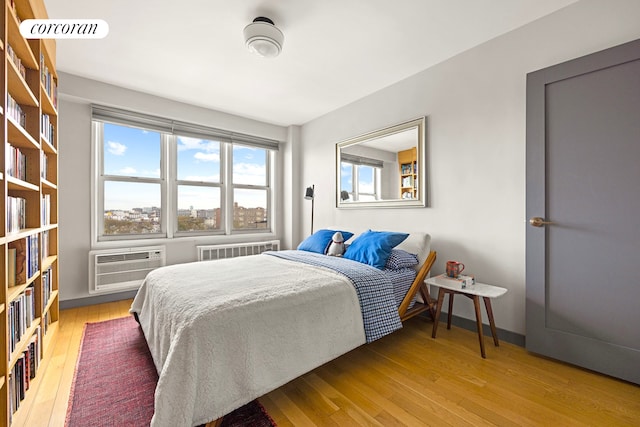 bedroom with radiator heating unit, a wall mounted air conditioner, wood finished floors, and baseboards
