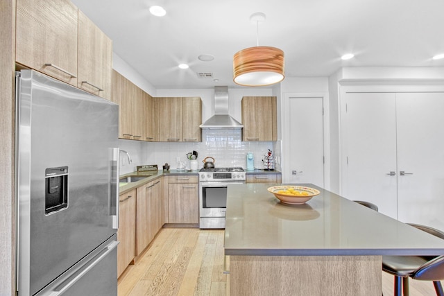 kitchen with appliances with stainless steel finishes, a sink, light brown cabinets, wall chimney exhaust hood, and light wood-type flooring