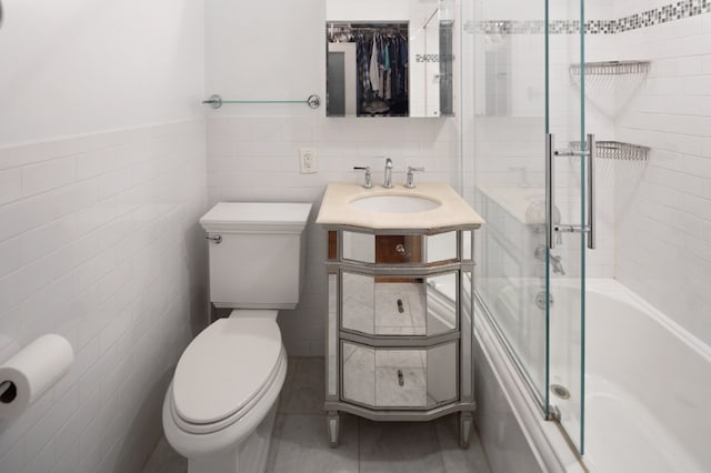full bathroom featuring tile walls, toilet, bath / shower combo with glass door, vanity, and a wainscoted wall