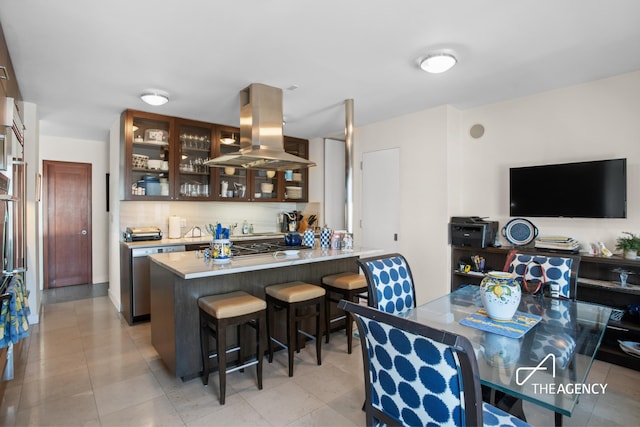 kitchen featuring island exhaust hood, a breakfast bar area, backsplash, a peninsula, and glass insert cabinets
