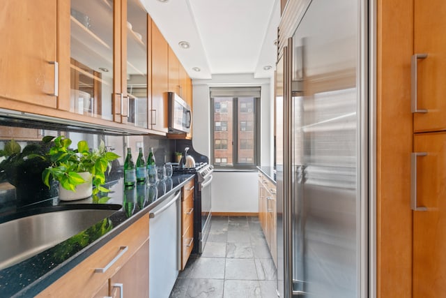 kitchen featuring stainless steel appliances, stone tile flooring, glass insert cabinets, baseboards, and brown cabinetry