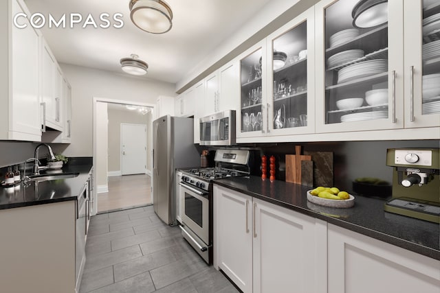 kitchen with appliances with stainless steel finishes, a sink, white cabinetry, dark countertops, and glass insert cabinets