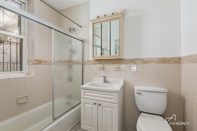 full bath featuring tile walls, combined bath / shower with glass door, toilet, vanity, and a wainscoted wall