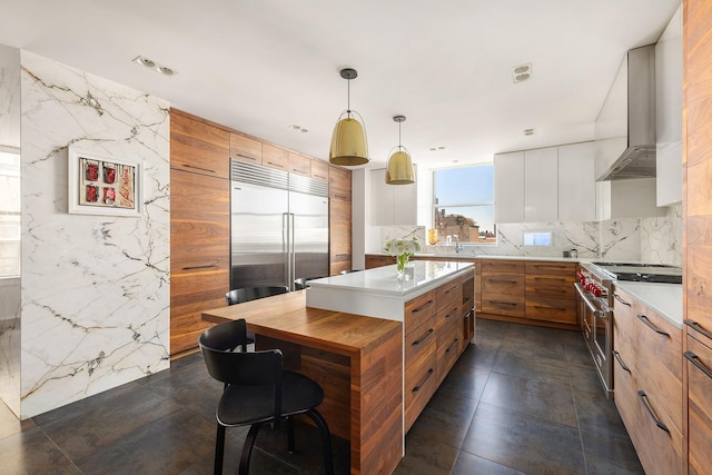kitchen with high end appliances, brown cabinetry, tasteful backsplash, a center island, and wall chimney range hood