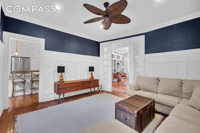 living room featuring a ceiling fan, crown molding, wainscoting, wood finished floors, and a decorative wall