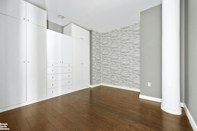 unfurnished bedroom featuring an accent wall, wallpapered walls, baseboards, visible vents, and dark wood-style floors