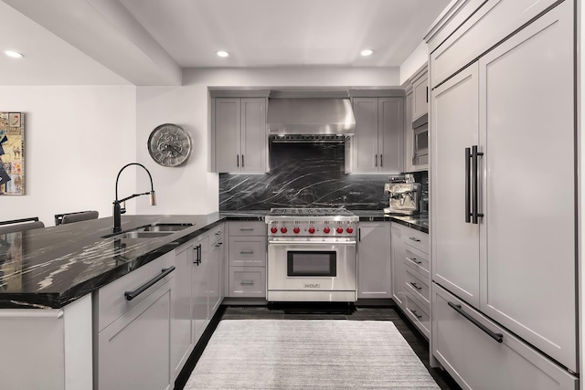 kitchen with decorative backsplash, built in appliances, a peninsula, wall chimney range hood, and a sink