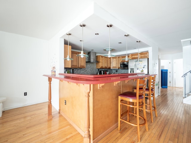 kitchen featuring wall chimney range hood, decorative backsplash, fridge with ice dispenser, stainless steel microwave, and light wood-style flooring