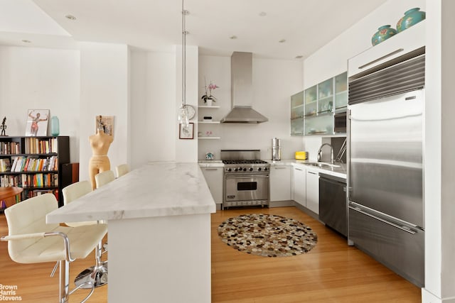 kitchen featuring high quality appliances, wall chimney exhaust hood, a peninsula, a sink, and light wood-style floors