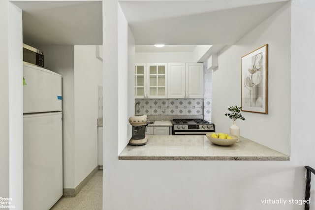 kitchen with white cabinets, stainless steel gas stove, freestanding refrigerator, backsplash, and glass insert cabinets