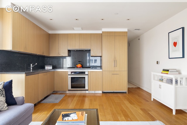 kitchen with decorative backsplash, oven, light wood-style floors, ventilation hood, and a sink