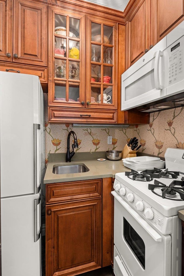 kitchen with brown cabinets, a sink, white appliances, light countertops, and glass insert cabinets