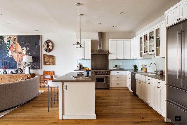 kitchen with wood finished floors, high end appliances, dark countertops, wall chimney exhaust hood, and a sink
