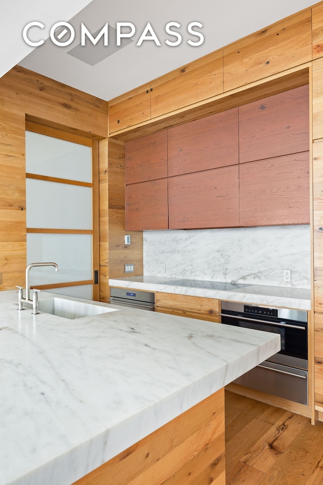 kitchen with light wood-type flooring, wood walls, a sink, light stone counters, and stainless steel oven