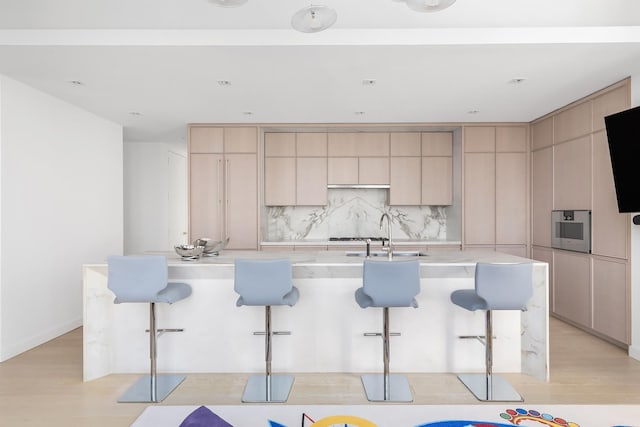 kitchen with tasteful backsplash, a sink, an island with sink, light wood-style flooring, and a kitchen bar