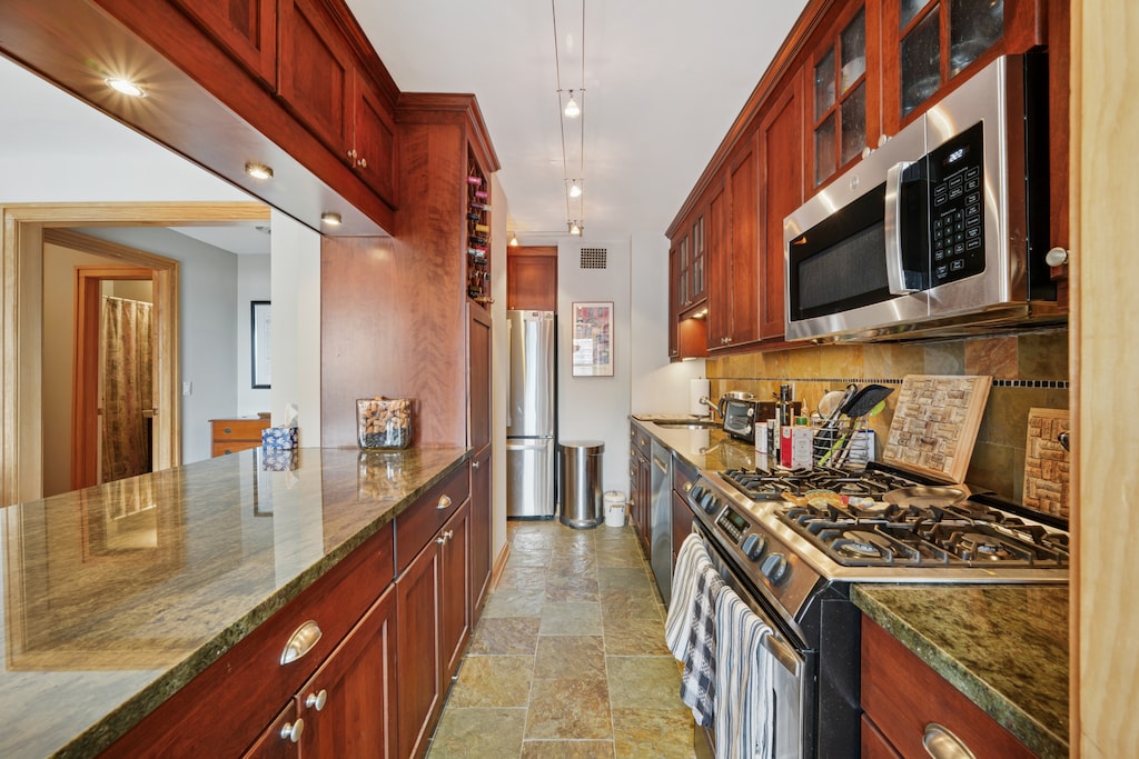 160 W End Ave Unit 24Gh, New York City, NY 10023 - kitchen with appliances with stainless steel finishes, stone finish floors, decorative backsplash, dark stone counters, and glass insert cabinets