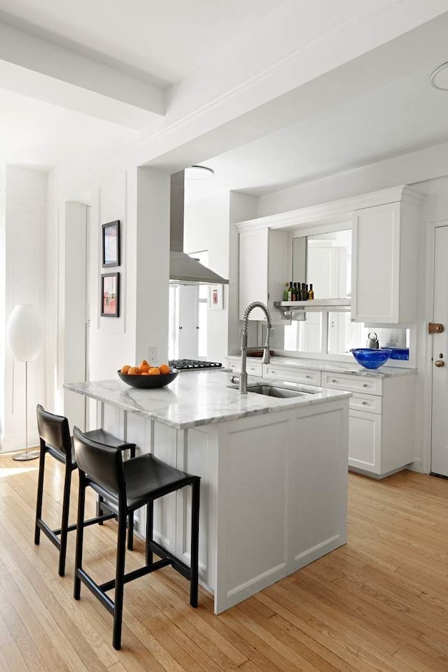 kitchen with wall chimney exhaust hood, white cabinetry, a sink, light stone counters, and light wood-style flooring