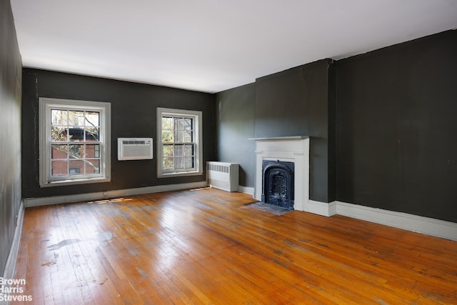 unfurnished living room featuring an AC wall unit, radiator, baseboards, a fireplace with flush hearth, and hardwood / wood-style flooring