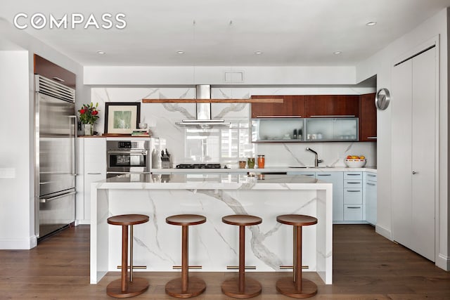 kitchen with dark wood-style flooring, ventilation hood, appliances with stainless steel finishes, and a breakfast bar area