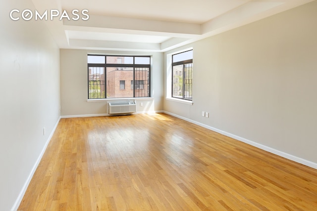 empty room featuring a wall unit AC, a tray ceiling, light wood-type flooring, and baseboards
