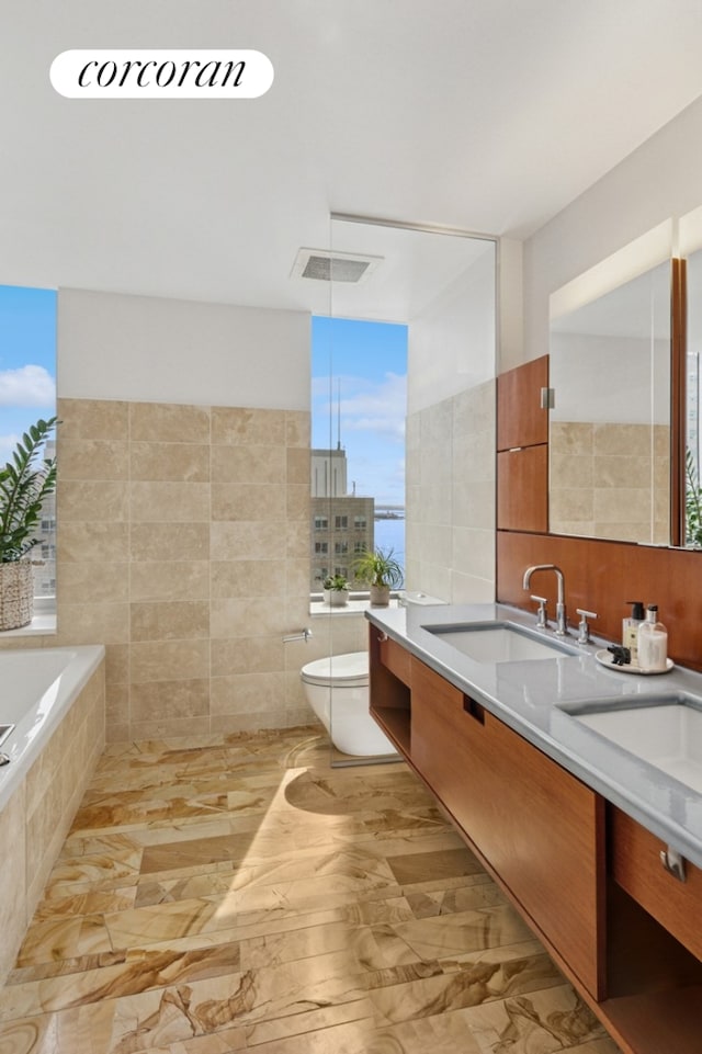 2 River Ter Unit 23C, New York City, NY 10282 - bathroom featuring tile walls, toilet, double vanity, a relaxing tiled tub, and marble finish flooring