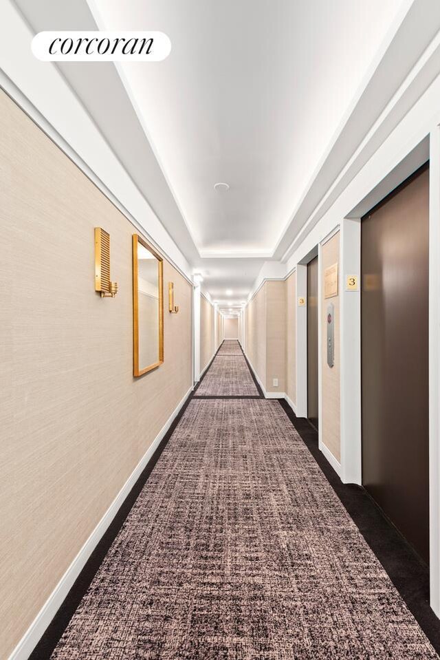 corridor featuring dark colored carpet, elevator, and baseboards