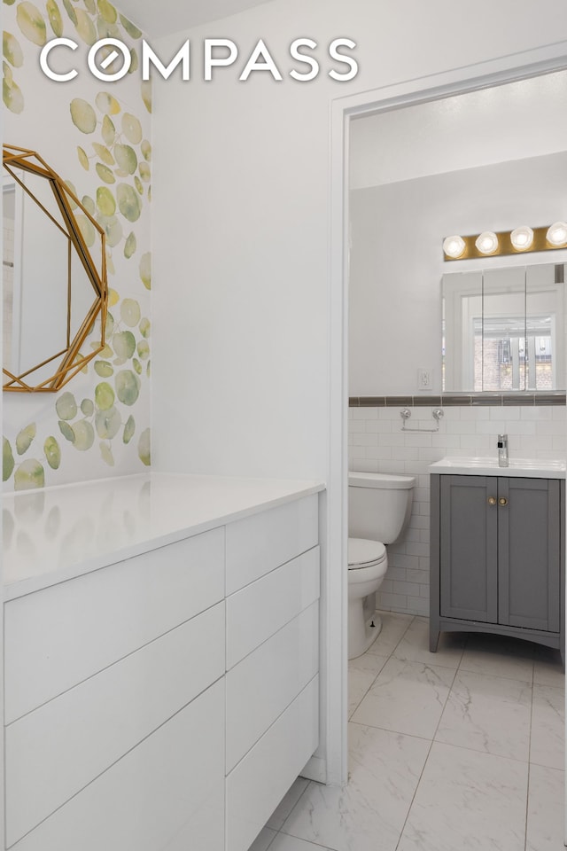 bathroom featuring marble look tiles, toilet, vanity, tile walls, and a wainscoted wall
